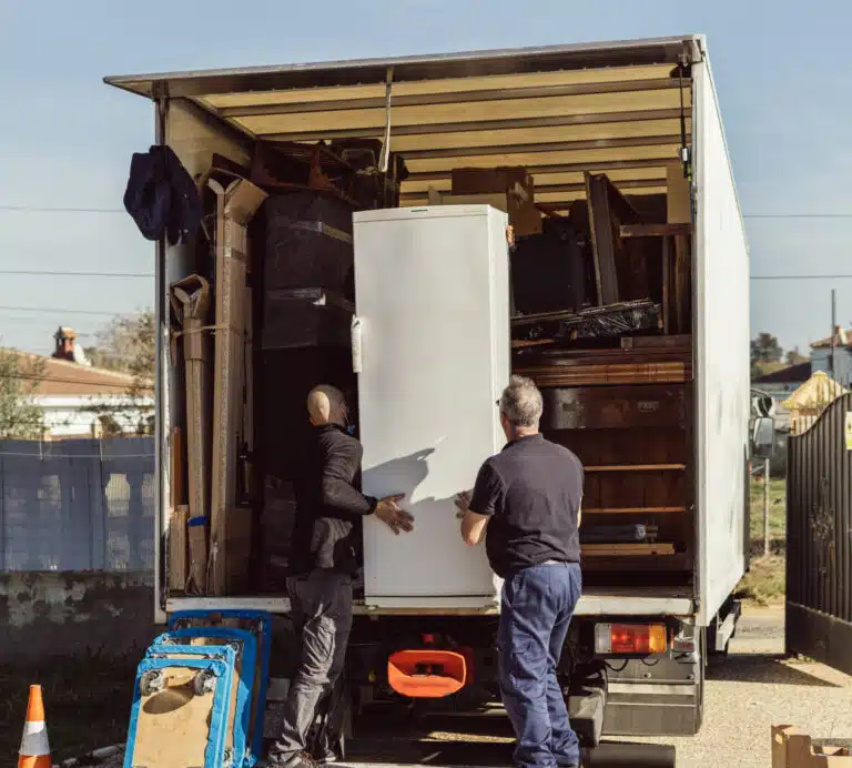 Two local movers carefully load a white refrigerator into a moving truck, amidst various other household items and furniture. The sunny outdoor setting suggests a residential move.