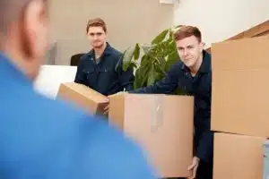 Two friendly local movers in blue uniforms carefully carry and stack cardboard boxes during a house move. They smile at the camera, showcasing their professional and efficient service.