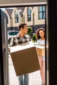 Man with a van Cootehill by Local Movers 27 Happy young couple carrying moving boxes into their new home, smiling and looking at each other. They are using local movers for their relocation. The boxes are brown cardboard, and they stand on a paved walkway in front of a modern house.