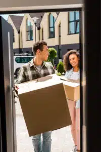 Happy young couple carrying moving boxes into their new home, smiling and looking at each other. They are using local movers for their relocation. The boxes are brown cardboard, and they stand on a paved walkway in front of a modern house.
