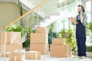 Man with a van Cootehill by Local Movers 26 A focused mover in blue overalls uses his phone while checking a clipboard, surrounded by stacked cardboard boxes in a bright, plant-filled building. This local moving company professional ensures a smooth relocation.