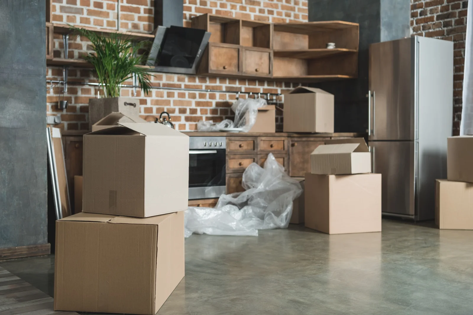 Local Movers Ireland 21 Cardboard boxes stacked in a partially unpacked kitchen, indicating a recent move. The room features exposed brick, wooden cabinets, and stainless steel appliances. Bubble wrap and packing materials are scattered on the floor, suggesting ongoing relocation efforts by local movers.