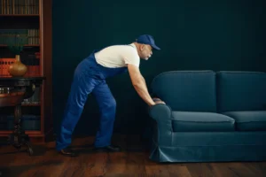 Man with a van Cootehill by Local Movers 24 A senior male mover in blue overalls and a blue cap carefully pushes a teal sofa across a hardwood floor in a home. The room features dark wood furniture and a bookcase filled with books, highlighting the professional local moving services provided.