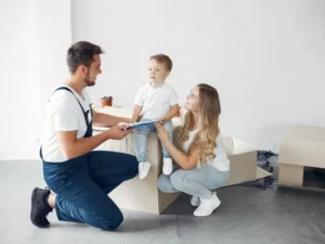 Man with a van Cootehill by Local Movers 23 Happy family with young child smiles at their local movers during a home move. The movers are kneeling, holding paperwork, while the family sits on a cardboard box in a new home setting.