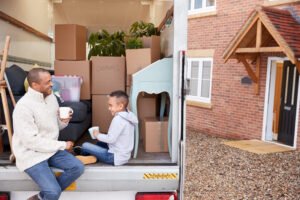 Man with a van Cootehill by Local Movers 17 A father and son enjoy a coffee break amidst moving boxes in the back of a moving truck. The happy family pauses during their move to a new home, showcasing the ease and care provided by local movers. "Cushions" is labeled on one box.