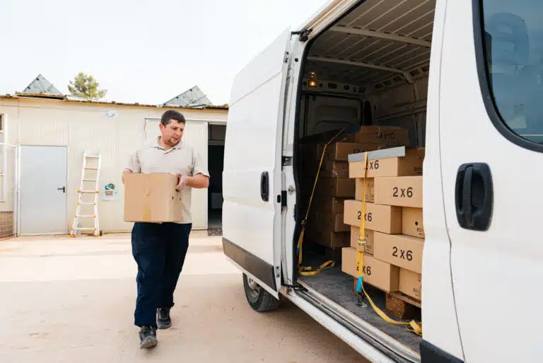 A delivery driver, wearing a beige polo shirt and dark pants, carefully carries a cardboard box from a white cargo van. The van is loaded with additional boxes marked "2x6," suggesting a local moving or delivery service.