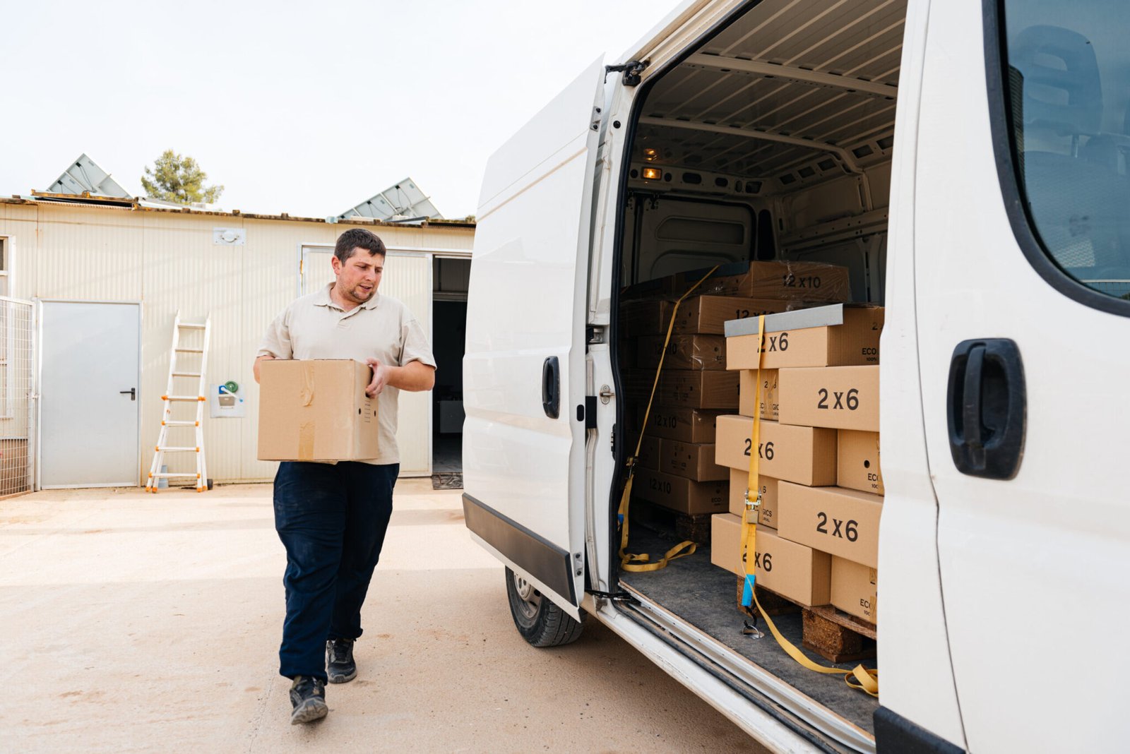 Local Movers Ireland 24 A delivery driver, wearing a beige polo shirt and dark pants, carefully carries a cardboard box from a white cargo van. The van is loaded with additional boxes marked "2x6," suggesting a local moving or delivery service.