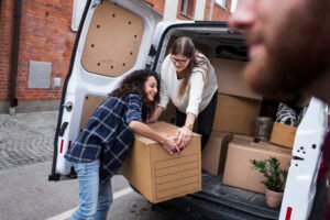 Man with a van Cootehill by Local Movers 19 Two women smile as they carefully load a large cardboard box into the back of a white moving van. One woman wears a cream-colored sweater, the other a blue and white plaid shirt. The van is packed with other boxes, suggesting a local move.