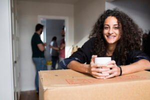 Man with a van Cootehill by Local Movers 16 Smiling young woman with curly brown hair sits on a cardboard box, holding a white mug, while local movers unpack in the background. She looks directly at the camera.
