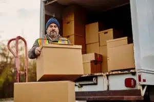 A focused local mover, wearing a safety vest and beanie, carefully carries a large cardboard box from a delivery truck, surrounded by numerous other packages. The scene suggests a busy moving day.