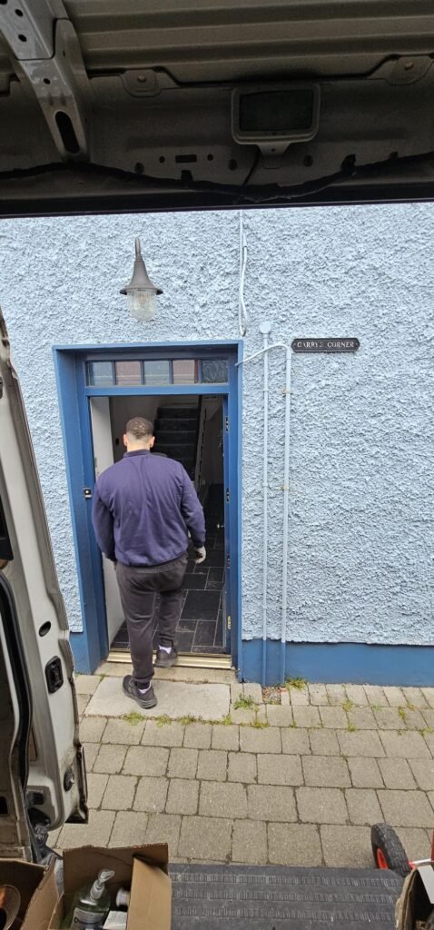 A mover in a navy jacket and black pants enters a blue doorway at "Carry's Corner," a house featured in an Irish Independent case study about local moving services.