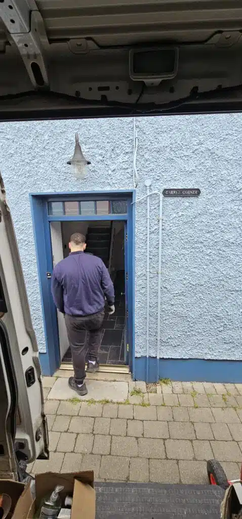 A mover in a navy jacket and black pants enters a blue doorway at "Carry's Corner," a house featured in an Irish Independent case study about local moving services.