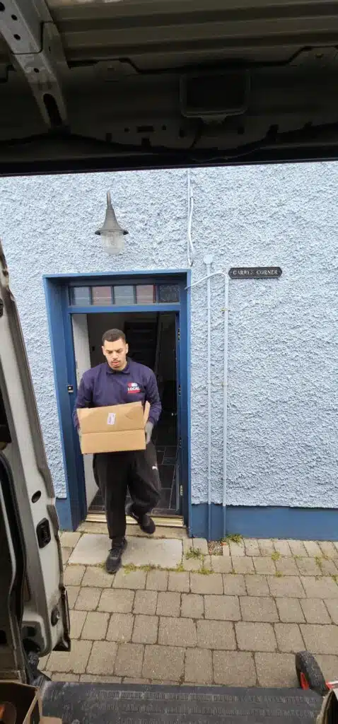 A local mover in a blue shirt with a "Local Movers" logo carries a cardboard box out of a blue doorway at "Carry Corner" house.