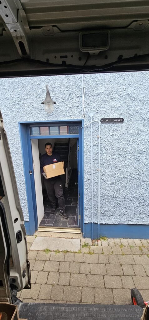 A local mover, wearing a dark blue uniform with "EMIGRÉ" visible, carries a cardboard box through the blue doorway of a house named "CAROL'S CORNER," as seen from inside a moving van.