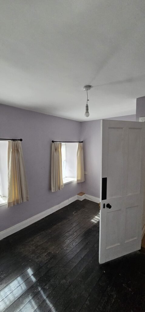 Interior of an empty, newly painted room with light purple walls, dark wooden floors, and two windows with cream curtains. This room is part of a house featured in an Irish Independent article about local movers.