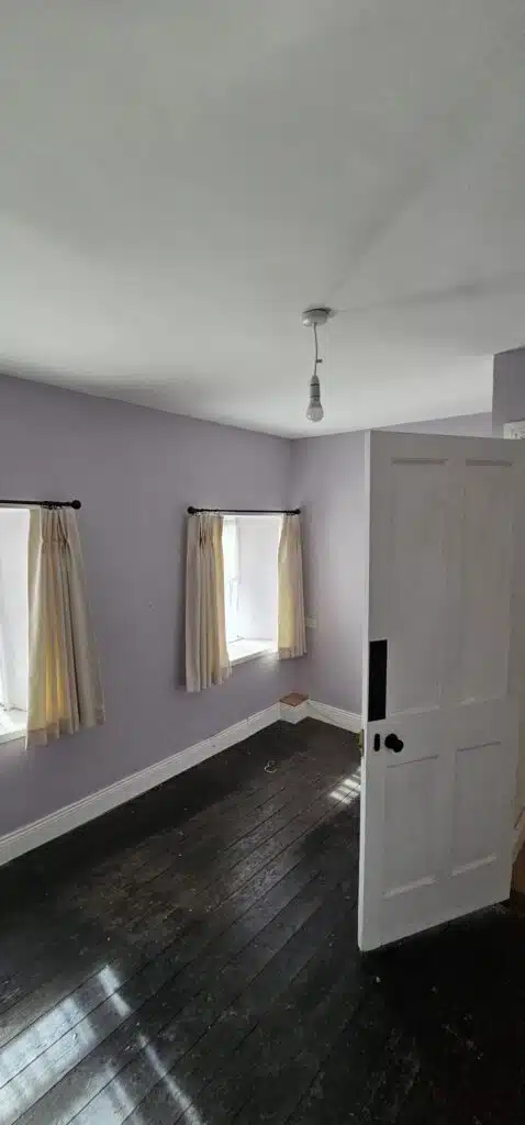 Interior of an empty, newly painted room with light purple walls, dark wooden floors, and two windows with cream curtains. This room is part of a house featured in an Irish Independent article about local movers.