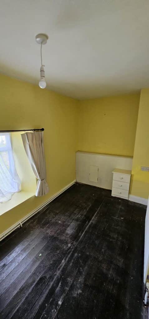 An empty, aged bedroom with dark wooden floors and bright yellow walls, featuring a window with sheer curtains and a white radiator.