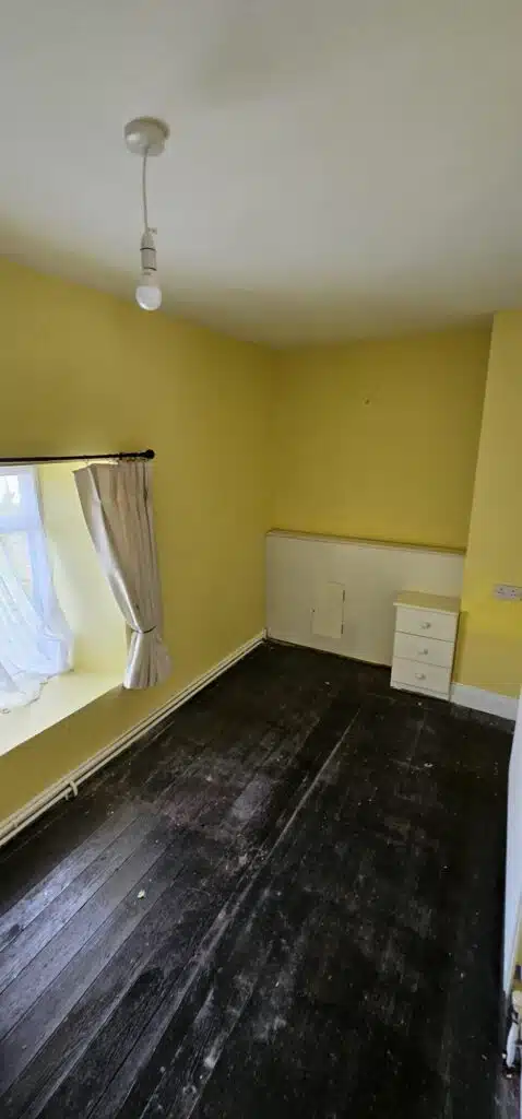 An empty, aged bedroom with dark wooden floors and bright yellow walls, featuring a window with sheer curtains and a white radiator.