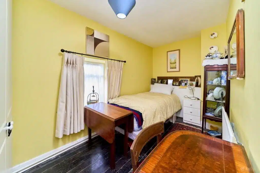 A bright yellow bedroom with a single bed, drop-leaf table, and antique display cabinet, potentially part of a house relocation featured in the Irish Independent.