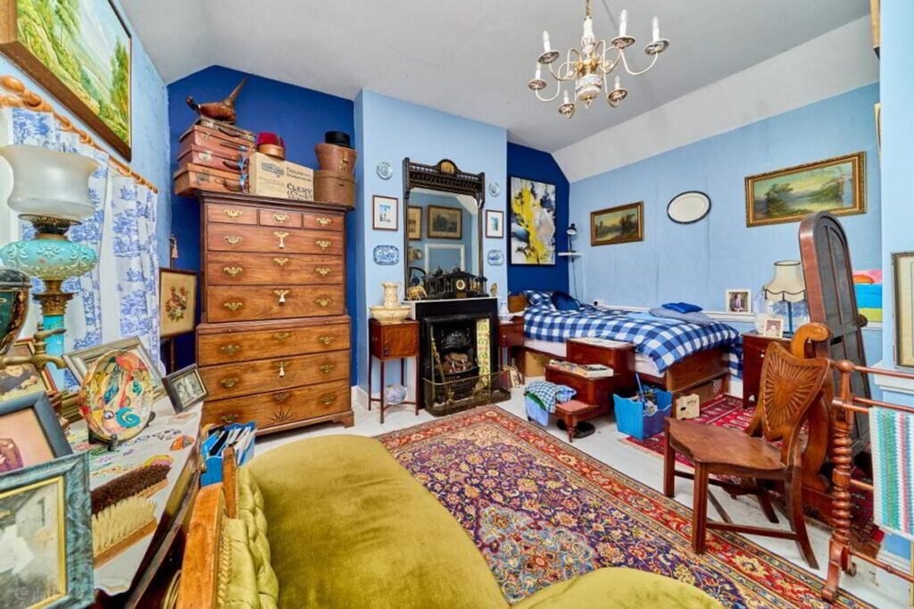 A cluttered, antique-filled bedroom with bright blue walls, a vintage chest of drawers, a patterned rug, and a four-poster bed, reminiscent of a home featured in a case study about local movers.