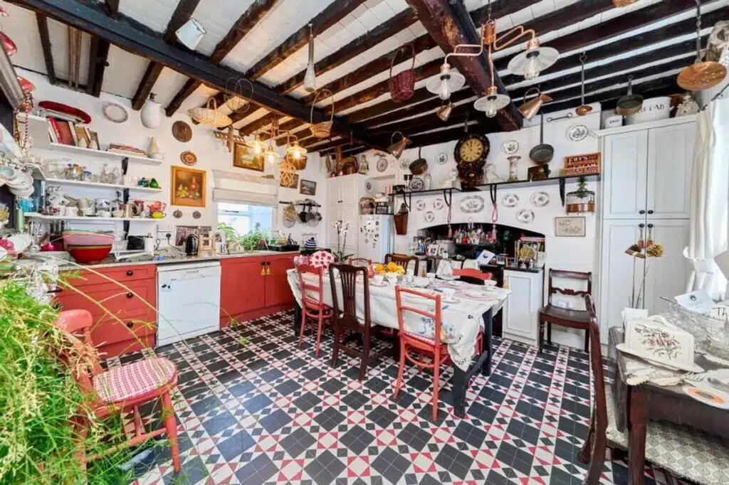 A charming, eclectic kitchen with a red and white tiled floor, a red island, and a dining table set for a meal. Exposed wooden beams and hanging baskets add rustic character, reminiscent of a cozy Irish home.