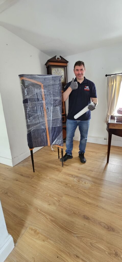 A Local Movers employee in a blue polo shirt and jeans gives a thumbs-up while holding packing supplies next to a tall, wrapped piece of furniture. A grandfather clock stands in the background.