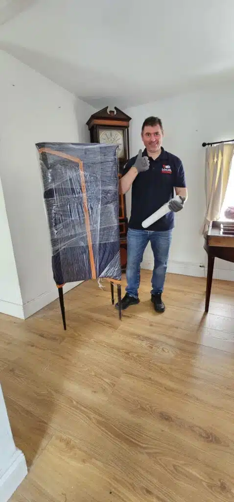 A Local Movers employee in a blue polo shirt and jeans gives a thumbs-up while holding packing supplies next to a tall, wrapped piece of furniture. A grandfather clock stands in the background.