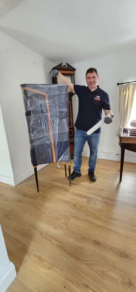 A man in a Local Movers uniform carefully wraps a dark blue armchair with plastic wrap, holding a roll of the material in his hand. He stands in a room with light wooden floors and a grandfather clock in the background.