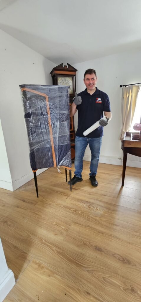 A Local Movers employee gives a thumbs-up while holding a roll of packing paper next to a grandfather clock and a piece of furniture wrapped in protective plastic, ready for relocation.
