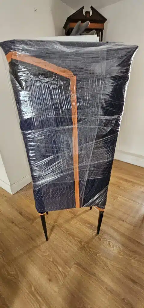 A navy blue upholstered chair, wrapped in protective padding and clear plastic, stands on a wooden floor, ready for relocation by local movers.