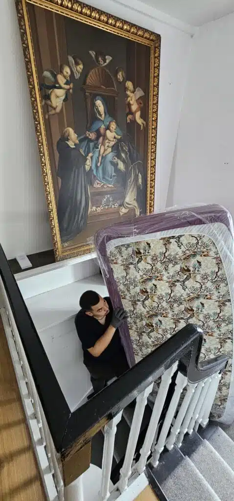 Movers carefully navigate a mattress down a staircase beside a large, ornate painting of the Madonna and Child, showcasing the challenges of relocating valuable art and furniture.