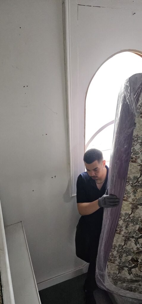 A mover in a navy shirt and grey gloves carefully carries a large, plastic-wrapped mattress up a stairwell, as seen in a case study on local movers.