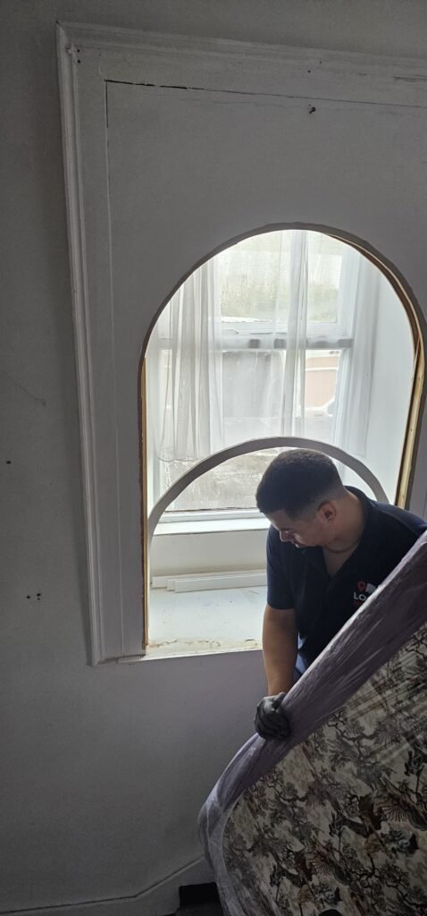 A mover in a dark blue shirt with a red and white logo carefully navigates a wrapped mattress through an arched doorway, showcasing the careful handling involved in relocating a house featured in the Irish Independent.