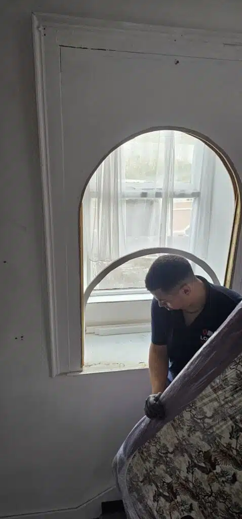 A mover in a dark blue shirt with a red and white logo carefully navigates a wrapped mattress through an arched doorway, showcasing the careful handling involved in relocating a house featured in the Irish Independent.