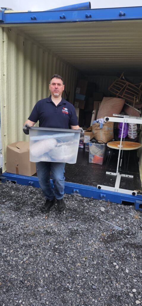 Man in a blue polo shirt and jeans, wearing gloves, holds a large clear plastic bin filled with items while standing in front of an open shipping container packed with boxes and furniture, ready for moving from Dublin to Sligo.