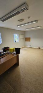 Empty office space with beige carpet, a wooden desk in the foreground, and fluorescent lighting overhead, indicating a commercial relocation.