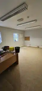 Empty office space with beige carpet, a wooden desk in the foreground, and fluorescent lighting overhead, indicating a commercial relocation.