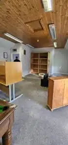 An empty office space featuring a wooden ceiling, a desk, a bookshelf, and a billiard table. A person stands near the door in the background. The room has a professional yet temporary feel with some furniture still in place.