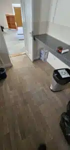 Newly installed wood-look vinyl flooring in a room with an open white door, a gray speckled countertop, and a recycling bin, suggesting a recent office move or renovation.