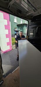 A man wearing gloves holds a large white desk in the back of a moving van parked on a street with a pink and white building and a black fence.