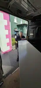 A man wearing gloves holds a large white desk in the back of a moving van parked on a street with a pink and white building and a black fence.