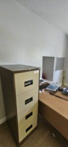 A beige and brown metal filing cabinet stands in an office setting next to a wooden desk with a computer monitor and papers, suggesting a commercial relocation.