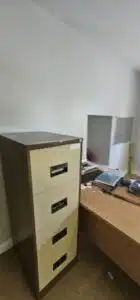 A beige and brown metal filing cabinet stands in an office setting next to a wooden desk with a computer monitor and papers, suggesting a commercial relocation.