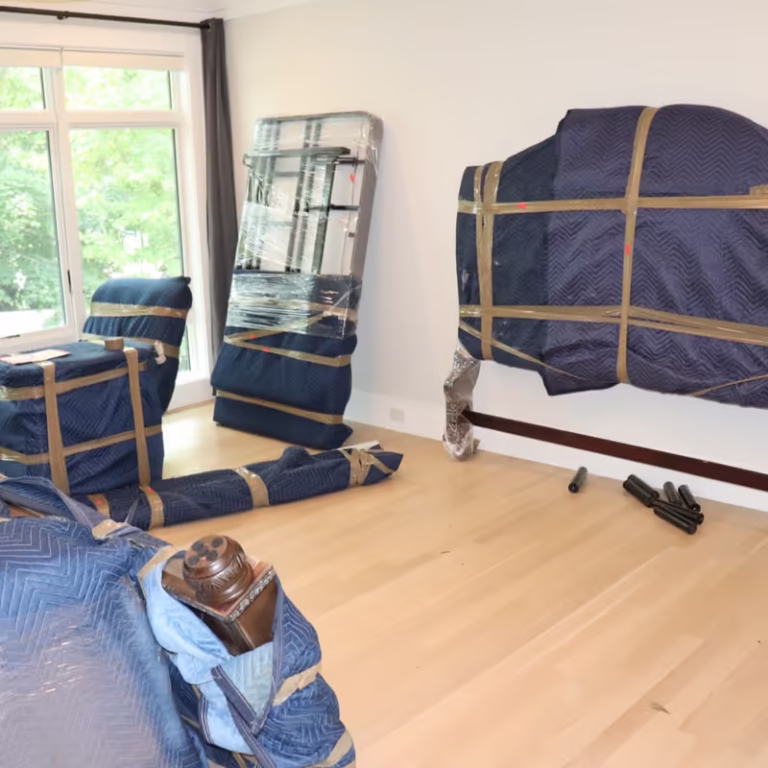 House removals in progress: Blue padded furniture, including a sofa, armchair, and bed frame, wrapped in plastic and tape, are staged in a bright room with large windows and light wood floors.