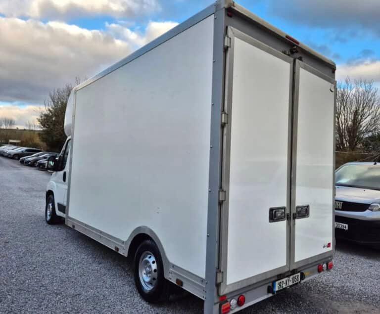 Rear view of a white box van with double rear doors, parked on a gravel lot with other vehicles and trees in the background. The van has a grey frame around the cargo area and license plate 192-KY-1058.