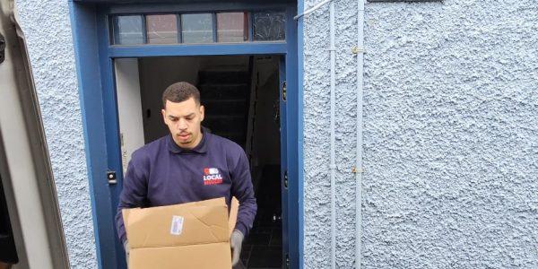 A local mover in a blue shirt with a "Local Movers" logo carries a cardboard box out of a blue doorway at "Carry Corner" house.