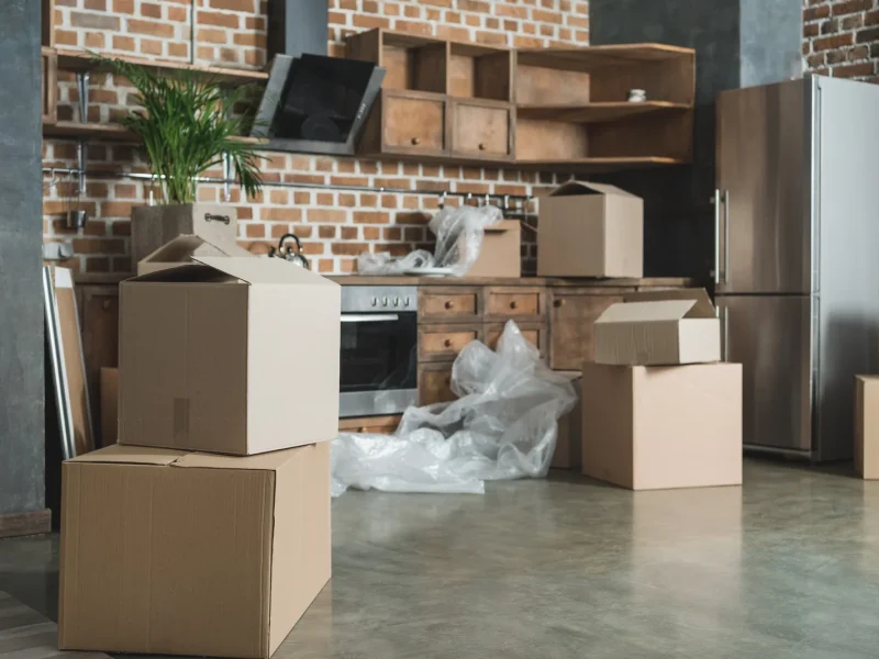 Cardboard boxes stacked in a partially unpacked kitchen, indicating a recent move. The room features exposed brick, wooden cabinets, and stainless steel appliances. Bubble wrap and packing materials are scattered on the floor, suggesting ongoing relocation efforts by local movers. Cardboard boxes stacked in a partially unpacked kitchen, indicating a recent move. The room features exposed brick, wooden cabinets, and stainless steel appliances. Bubble wrap and packing materials are scattered on the floor, suggesting ongoing relocation efforts by local movers.
