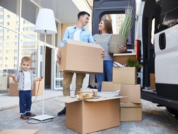 Happy family unloading boxes from a moving van outside their new home. A young boy playfully holds a lamp cord while his parents carry in large cardboard boxes. The scene is bright, cheerful, and depicts a successful move.